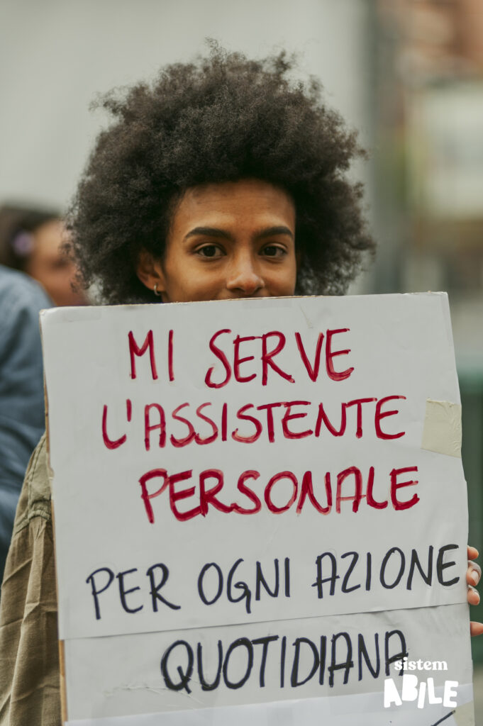 Una persona con un cartello che dice: "Mi serve l'assistenza personale per ogni azione quotidiana"